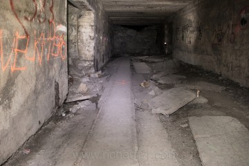 Rozlámaná podlaha a zbytky původního do betonu zapuštěného kolejiště. V zadní části fotografie první z velkých závalů v chodbě D.